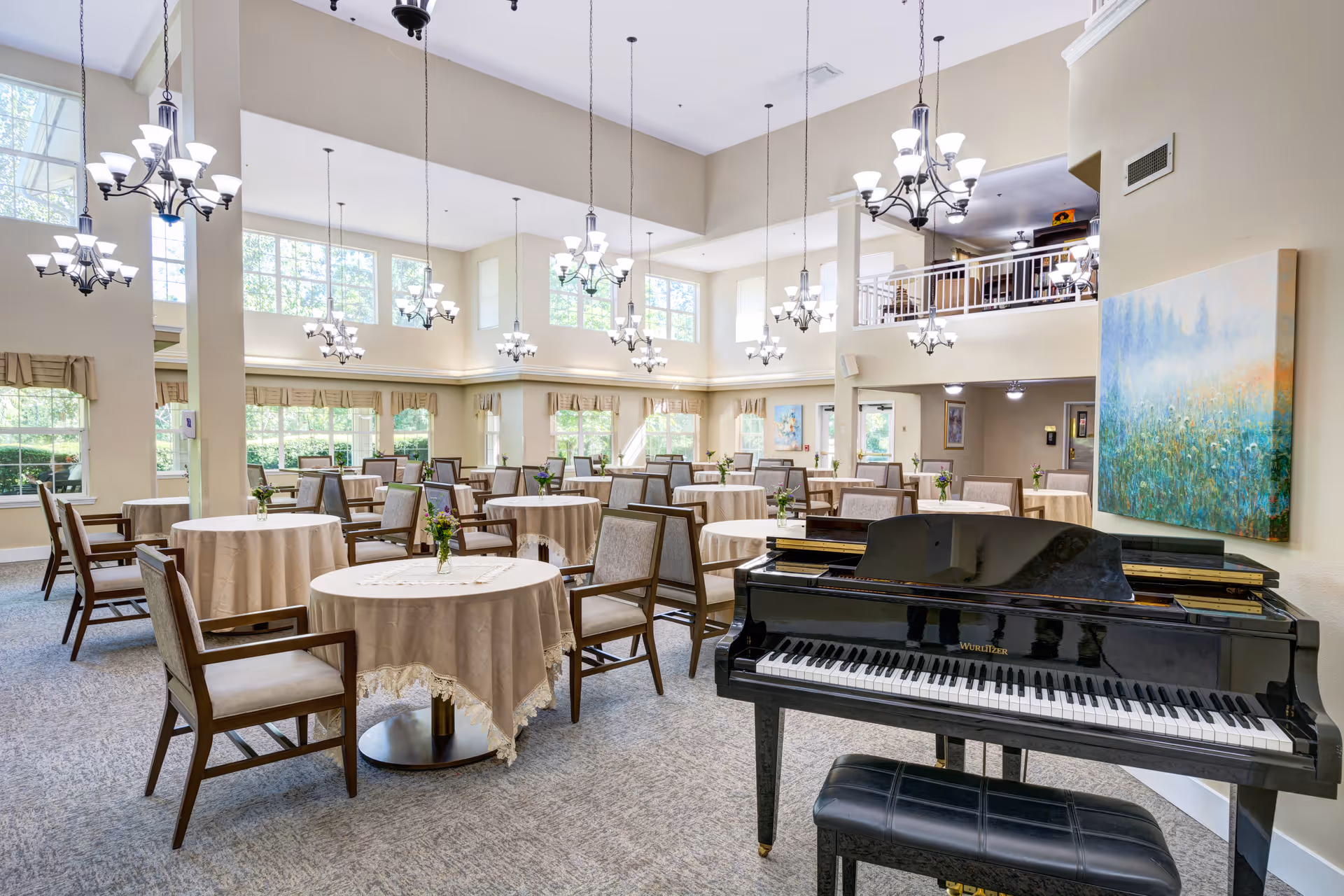 A spacious dining room with multiple round tables covered with beige tablecloths, each table adorned with a small flower arrangement. Wooden chairs with cushioned seats surround the tables. A black grand piano with a matching bench is positioned in the foreground. The room features large windows allowing natural light to fill the space, high ceilings with multiple hanging chandeliers, and light-colored walls decorated with artwork.