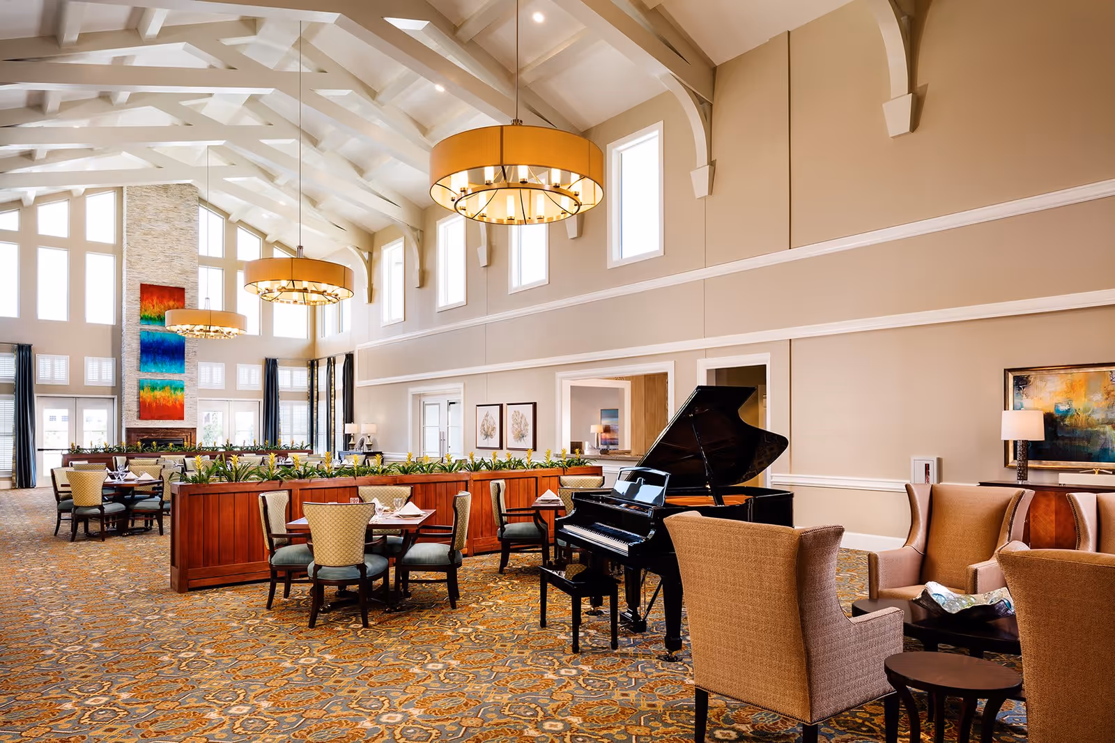 A spacious and elegant common area with high vaulted ceilings and large windows allowing natural light to fill the room. The room features a grand piano, several seating arrangements including armchairs and dining tables with chairs, decorative plants, and modern chandeliers hanging from the ceiling. The walls are painted beige with white trim, and there is colorful artwork on the walls and a tall stone fireplace in the background.