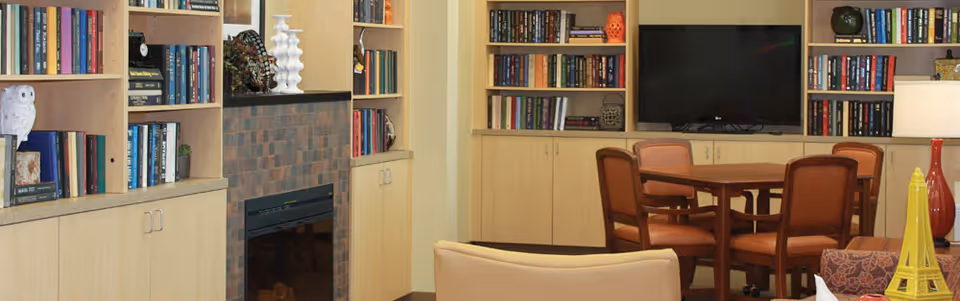 Interior common area in a senior living facility with built-in bookshelves, a brick fireplace, a television, and a round dining table with chairs.