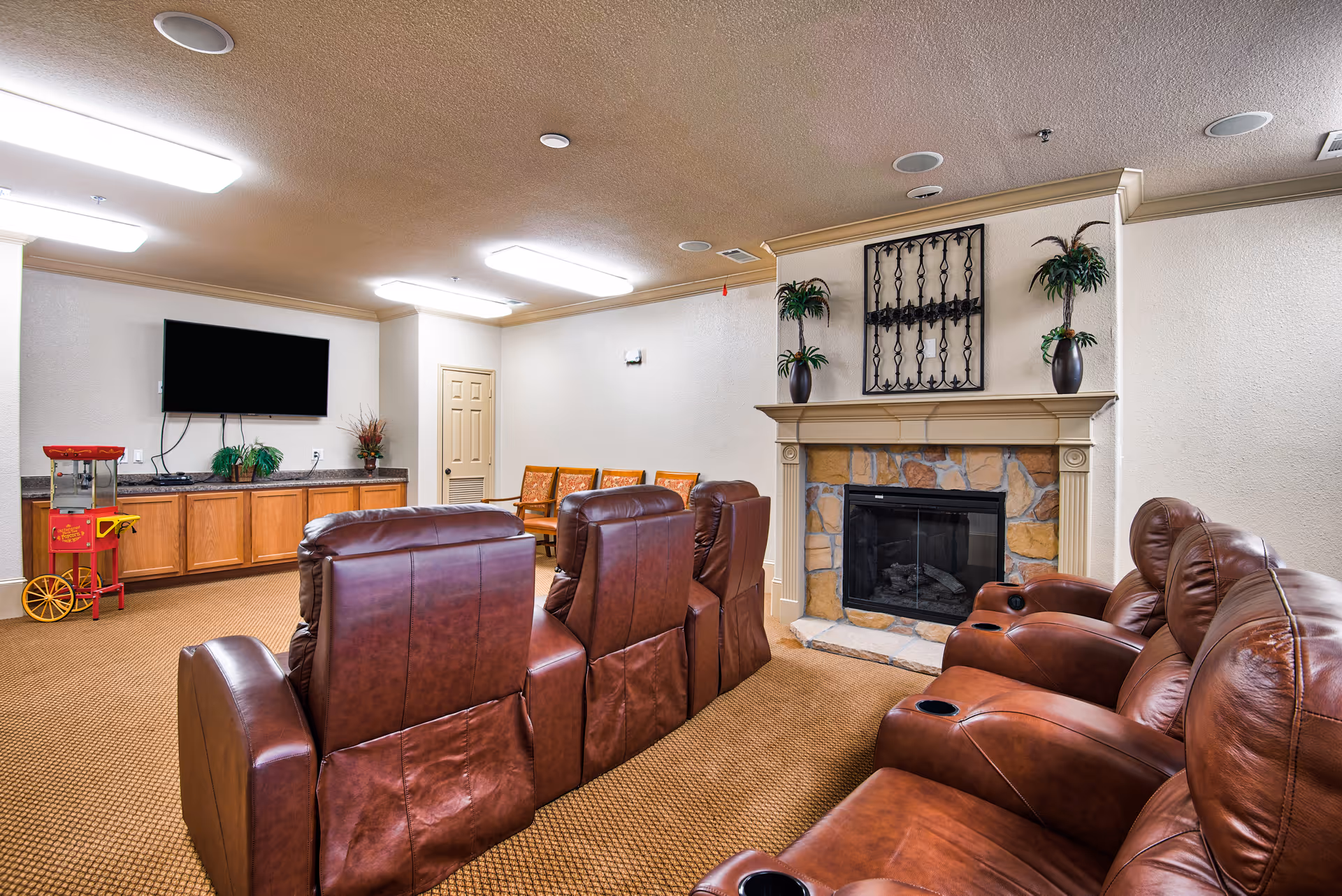 A cozy common area with brown leather recliner chairs arranged in rows facing a wall-mounted flat screen TV. There is a stone fireplace with a mantel decorated with two potted plants and a decorative metal wall hanging above it. A small popcorn machine is placed on a wooden cabinet along the wall, and several wooden chairs are lined up against the far wall. The room has beige walls and carpeted flooring.