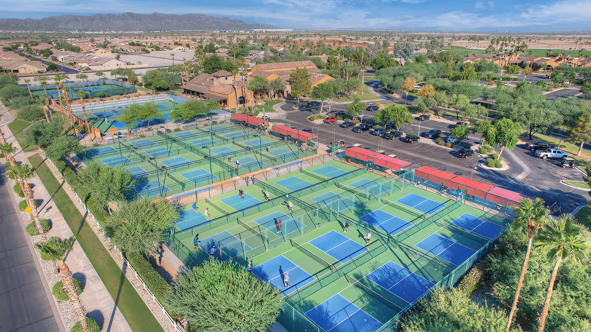 Aerial view of numerous fenced pickleball/tennis courts with players, adjacent parking, clubhouse buildings, and surrounding desert landscape.