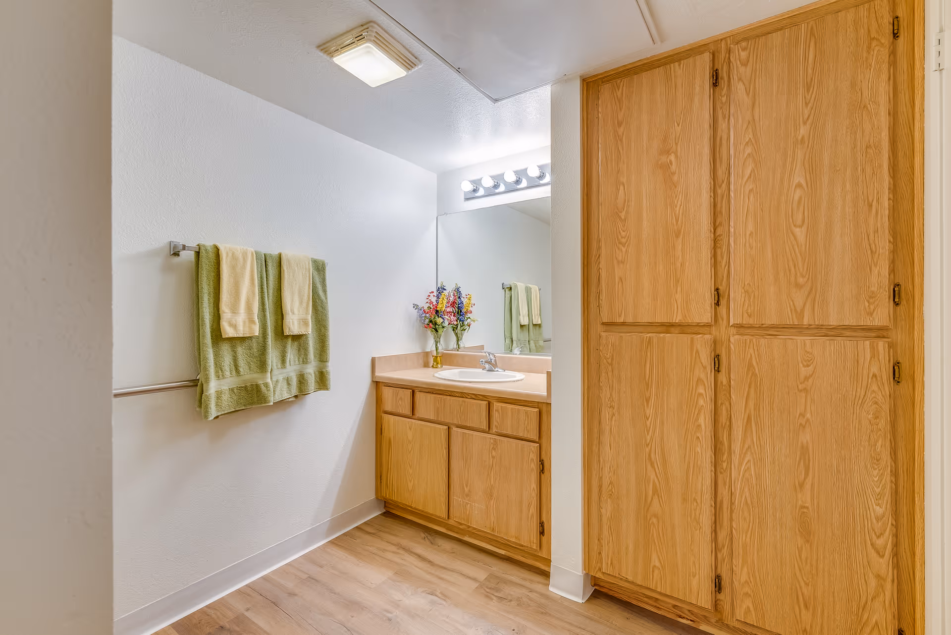 A bathroom interior featuring a wooden vanity with a sink and a large mirror above it. There are four light bulbs mounted above the mirror. On the left wall, two green towels with smaller beige towels on top hang on a towel rack. To the right of the vanity, there is a tall wooden cabinet with multiple doors. The floor is wood-style laminate, and the walls are painted white.