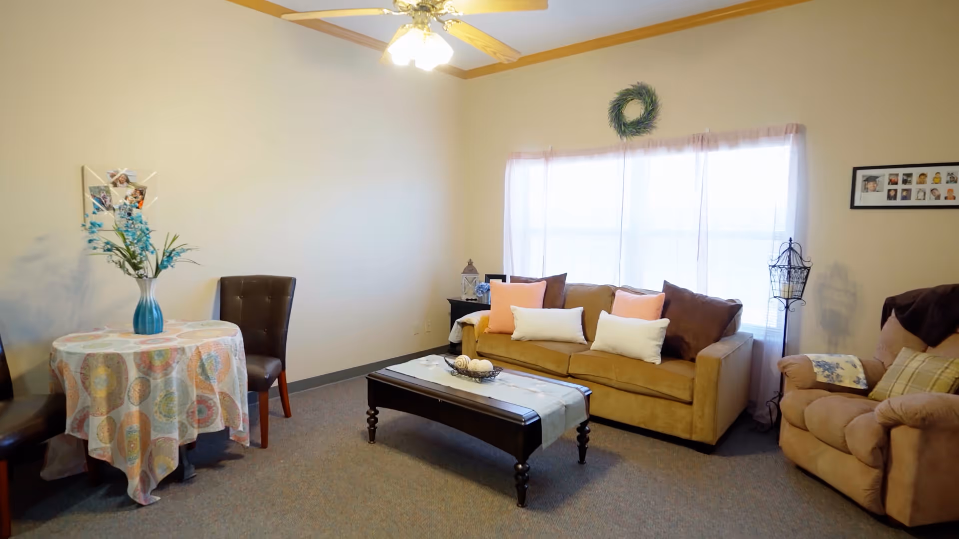 A cozy living room with a beige sofa adorned with multiple pillows, a cushioned armchair, a dark wooden coffee table with decorative items, and a small round table covered with a patterned tablecloth and a vase with flowers. The room has a ceiling fan with lights, a window with sheer curtains, and light-colored walls with minimal decorations.