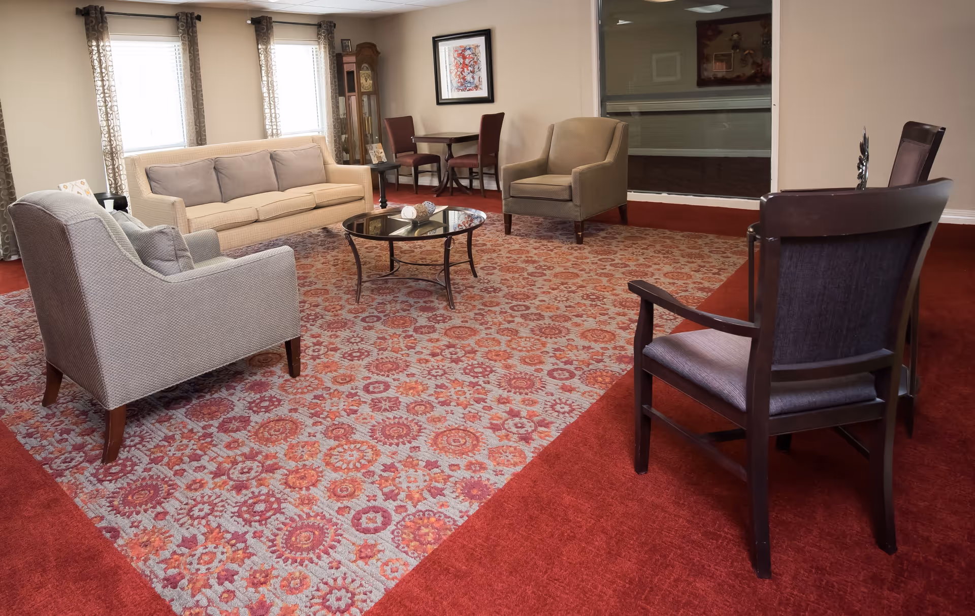 A cozy living room with a beige sofa, two armchairs, and two wooden chairs arranged around a glass coffee table on a patterned red and gray carpet. There are windows with patterned curtains, a grandfather clock, a small table with two chairs, and framed artwork on the wall.
