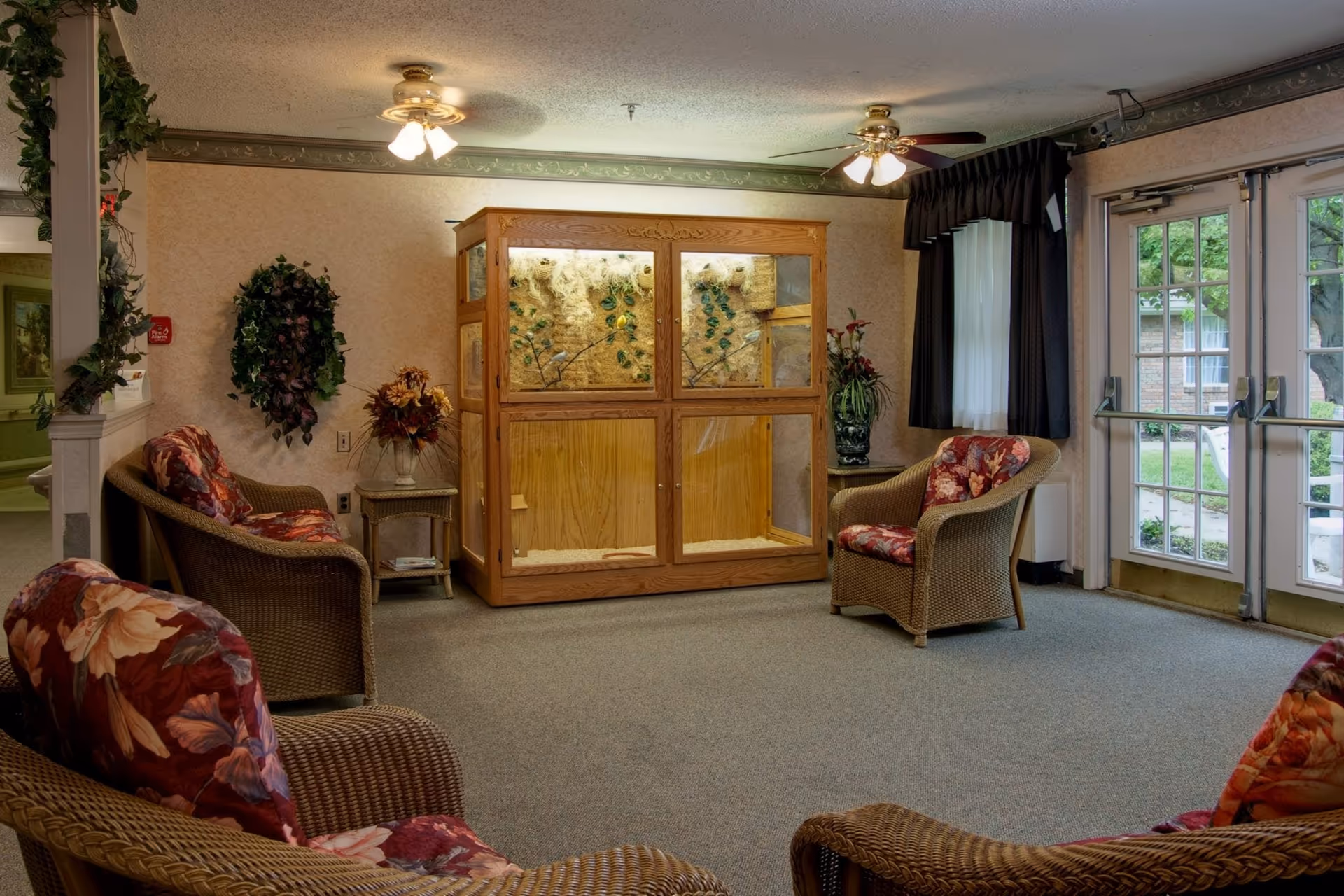 A cozy sitting area in a senior living facility with wicker chairs featuring floral cushions arranged around a large wooden enclosure with glass panels. The room has carpeted flooring, two ceiling fans with lights, decorative plants, and large glass doors leading outside.