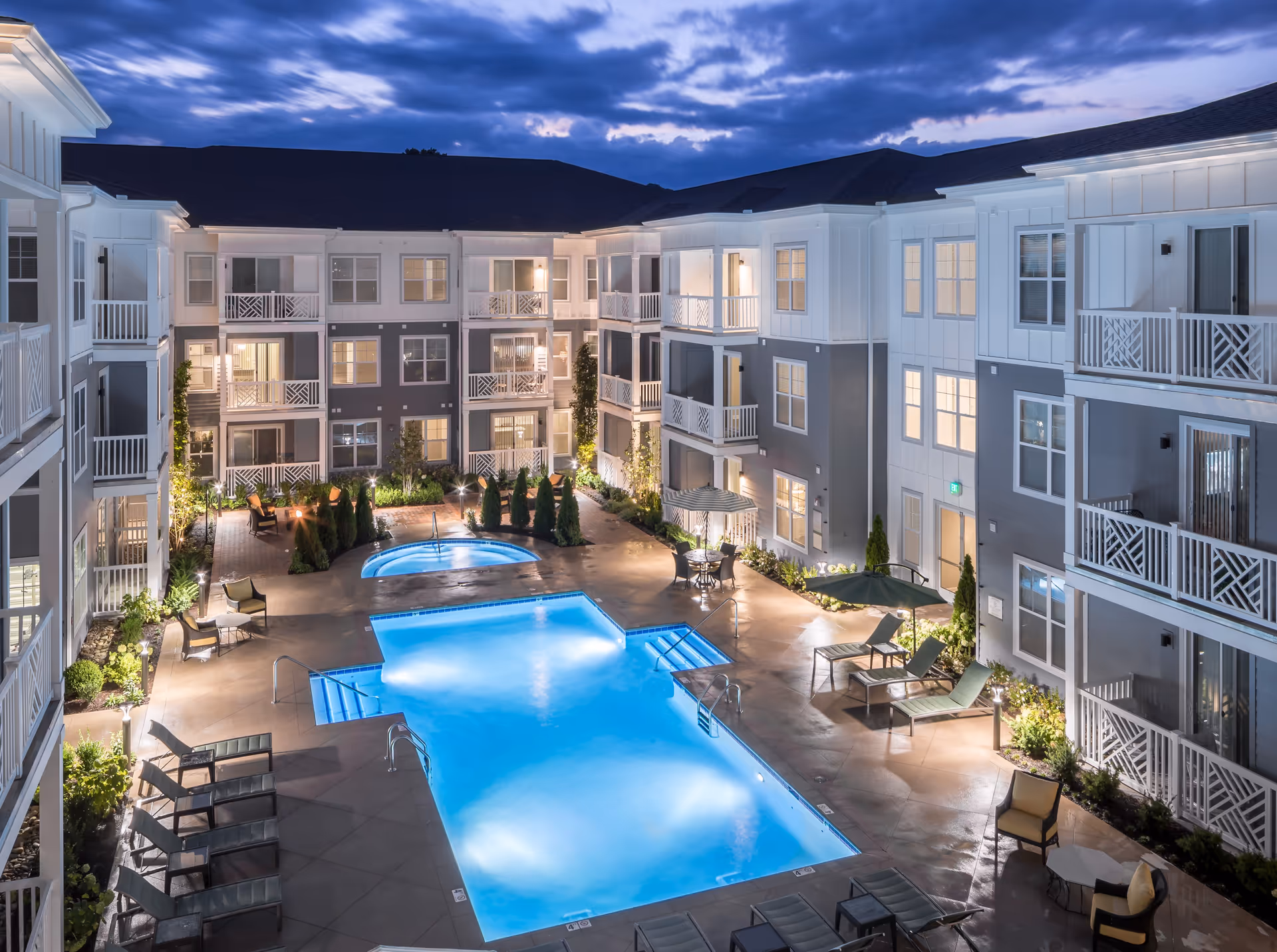 Night view of a modern senior living facility courtyard with a large illuminated swimming pool and a smaller hot tub. The courtyard is surrounded by a three-story building with balconies and windows lit from inside. There are lounge chairs, tables with umbrellas, and landscaped greenery around the pool area.