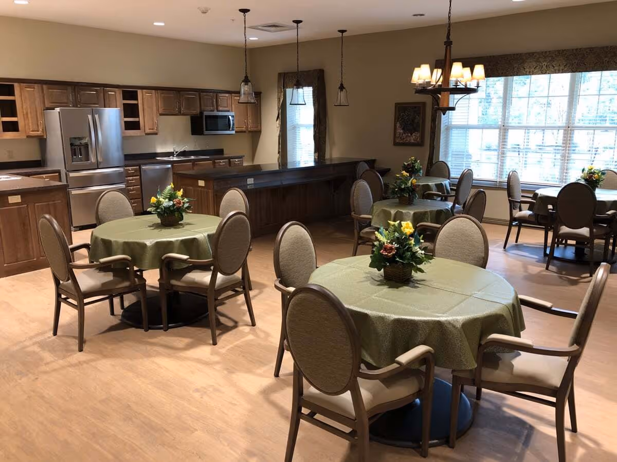 A dining area in a senior living facility featuring round tables covered with green tablecloths, each adorned with a floral centerpiece. The room includes several cushioned chairs with armrests around each table. In the background, there is a kitchen area with wooden cabinets, stainless steel refrigerator, dishwasher, and microwave. Large windows allow natural light to fill the space, and pendant lights hang from the ceiling.