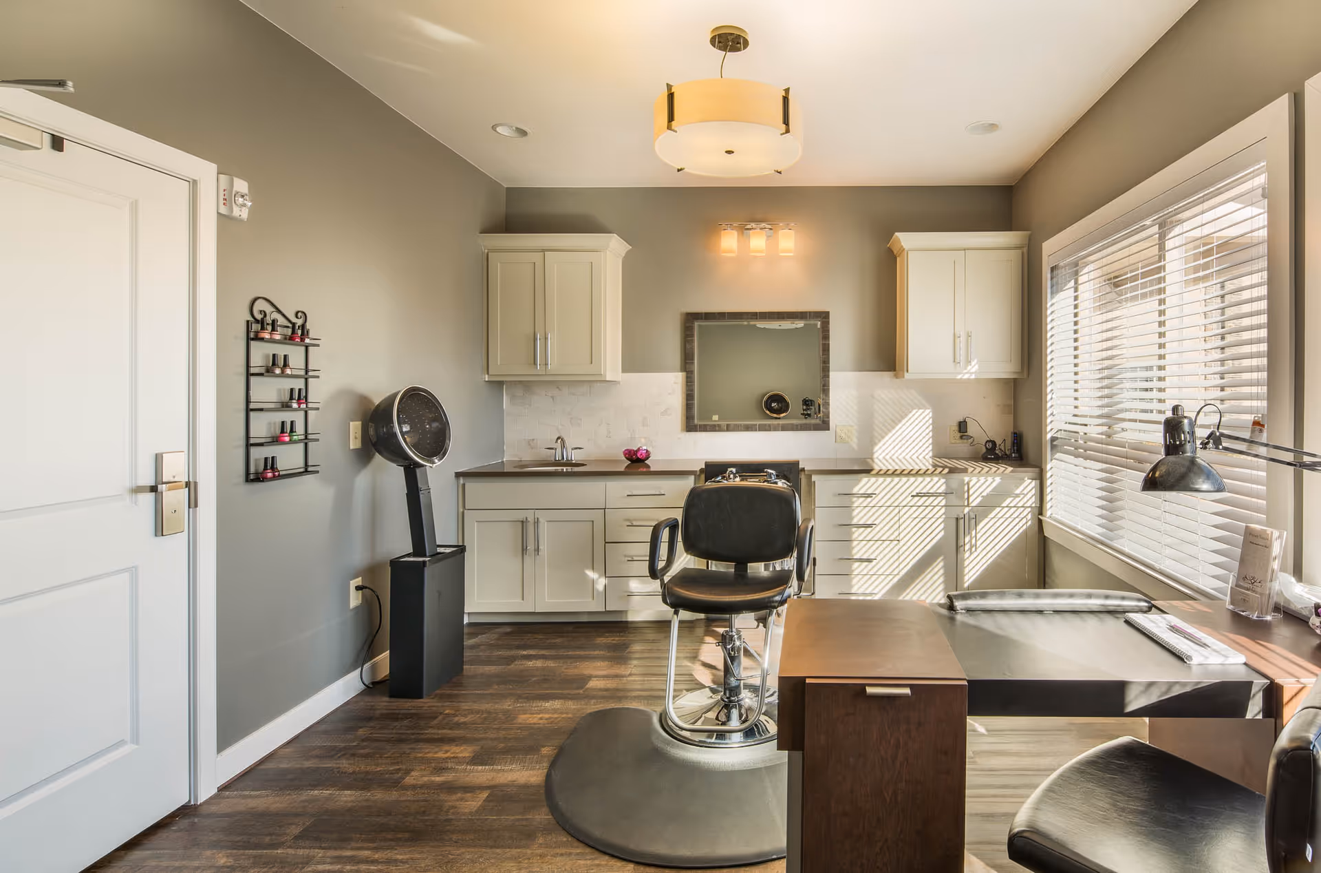 A bright and clean salon room with a black salon chair in front of a mirror mounted on a gray wall. The room features white cabinets, a hair dryer, a nail polish rack, a desk with a lamp, and large windows with white blinds letting in natural light.