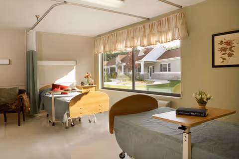 A bright and clean patient room with two hospital-style beds, one near a large window showing a view of a courtyard with buildings. The room has light-colored walls, a chair, a small table with a book and flowers, and a framed floral picture on the wall.