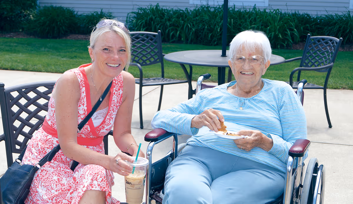 An elderly woman sitting in a wheelchair outdoors, smiling and holding a plate with a cookie. Next to her is a younger woman in a pink and white dress, also smiling and holding a cold drink with a straw. They are seated on a patio with metal chairs and tables, with green grass and plants in the background.