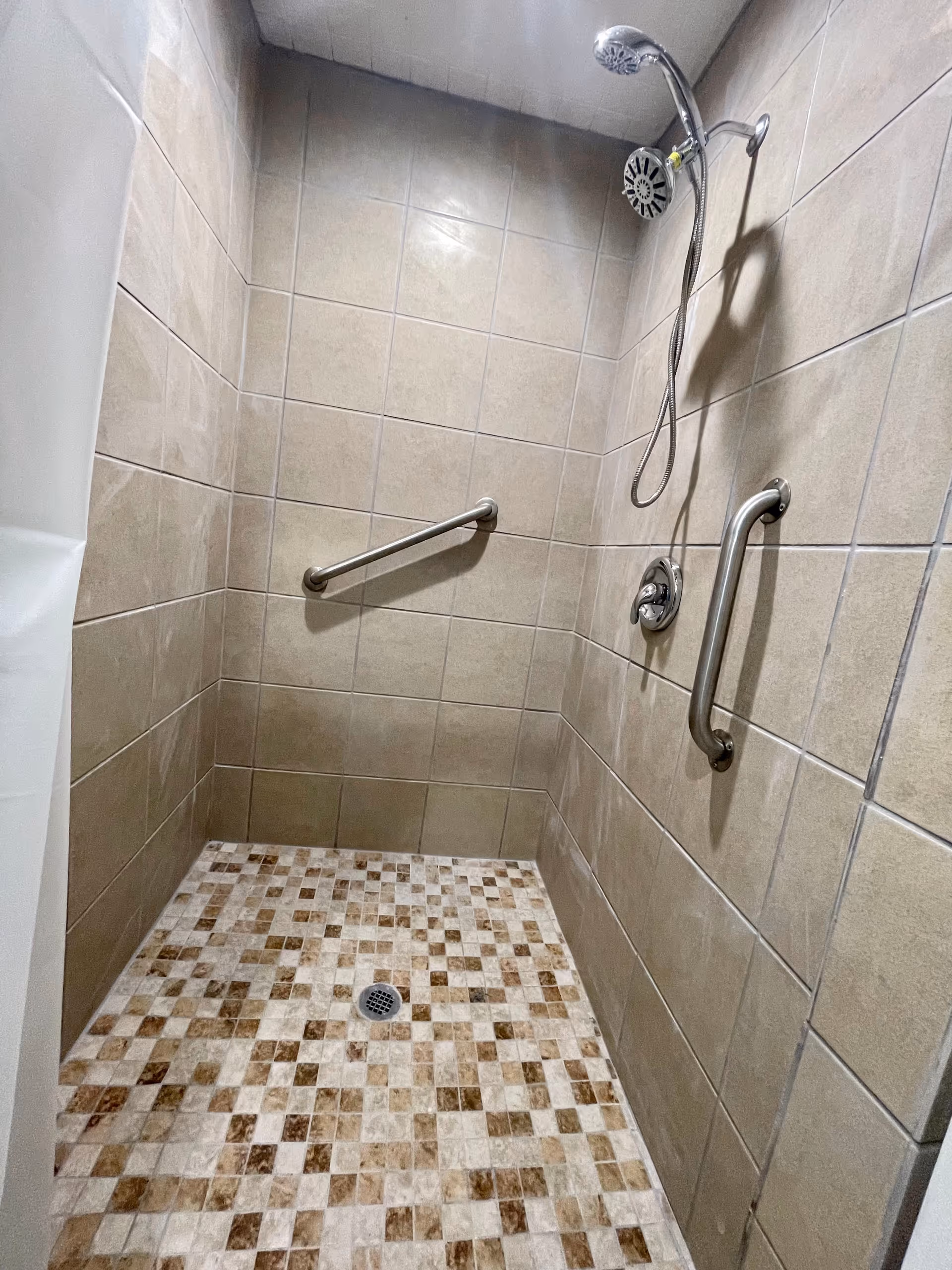 A tiled walk-in shower with beige wall tiles and a mosaic tile floor in shades of brown and cream. The shower has a chrome showerhead, two stainless steel grab bars on the walls, and a drain in the center of the floor.