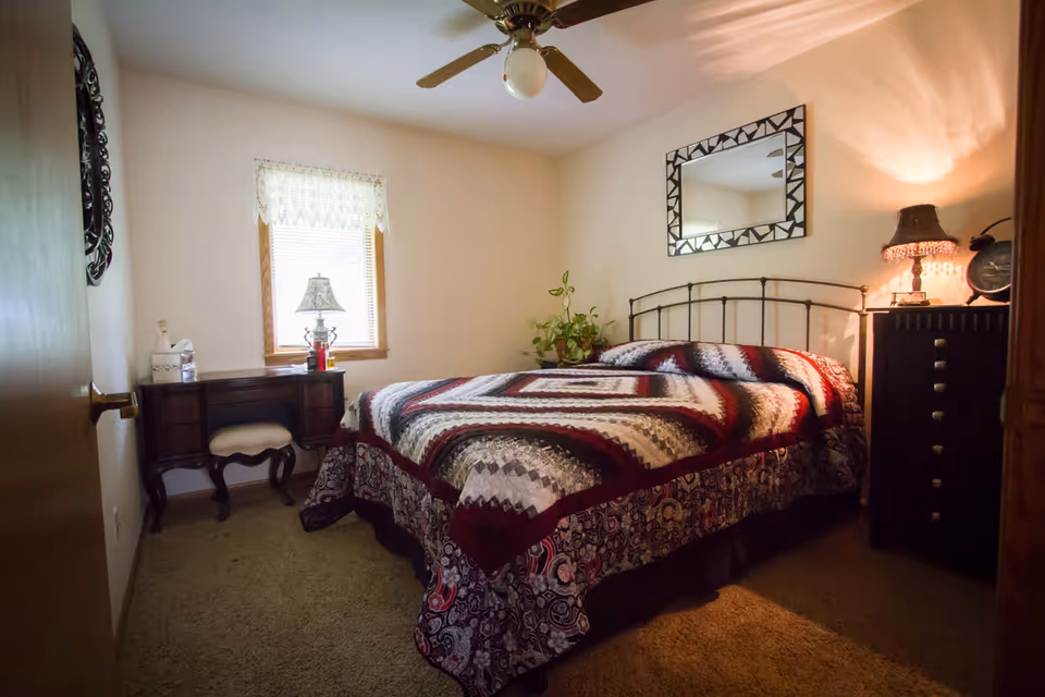A cozy bedroom with a bed covered in a patterned quilt, a decorative mirror above the bed, a bedside table with a lamp and clock, a small desk with a lamp and tissue box near a window with lace curtains, and a ceiling fan overhead.