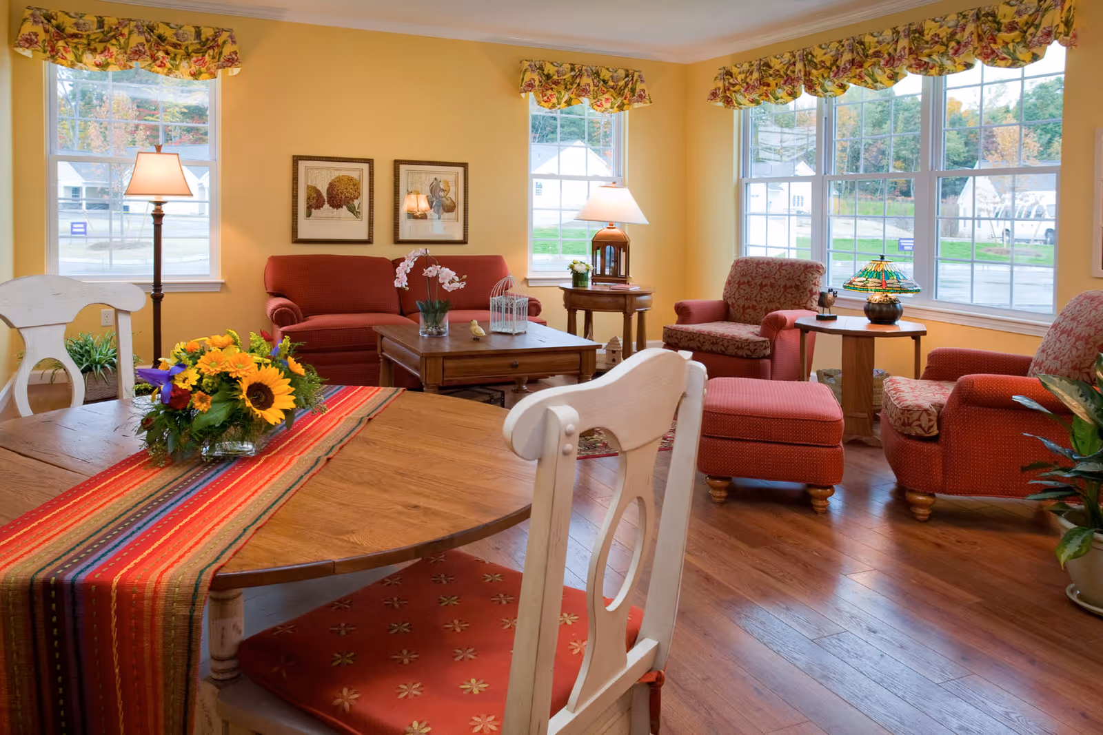 A cozy living room with yellow walls and large windows letting in natural light. The room features red upholstered armchairs and a matching ottoman, a red sofa, wooden coffee and side tables with lamps, and a wooden dining table with white chairs. A colorful striped table runner and a vase of sunflowers decorate the dining table. Floral valances adorn the windows.
