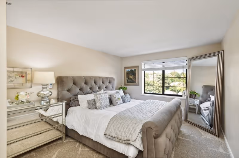 A bright and cozy bedroom featuring a large tufted grey upholstered bed with white and grey bedding and decorative pillows. To the left of the bed is a mirrored nightstand with a silver lamp and floral arrangement. A large window with white blinds lets in natural light, and a framed picture hangs on the wall next to the window. A tall, framed mirror leans against the wall on the right side of the room, reflecting part of the bed and nightstand. The room has beige walls and carpeted flooring.