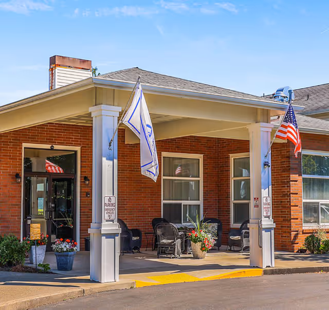 Brick assisted-living facility entrance with a covered porch, chairs, flags, and planters.
