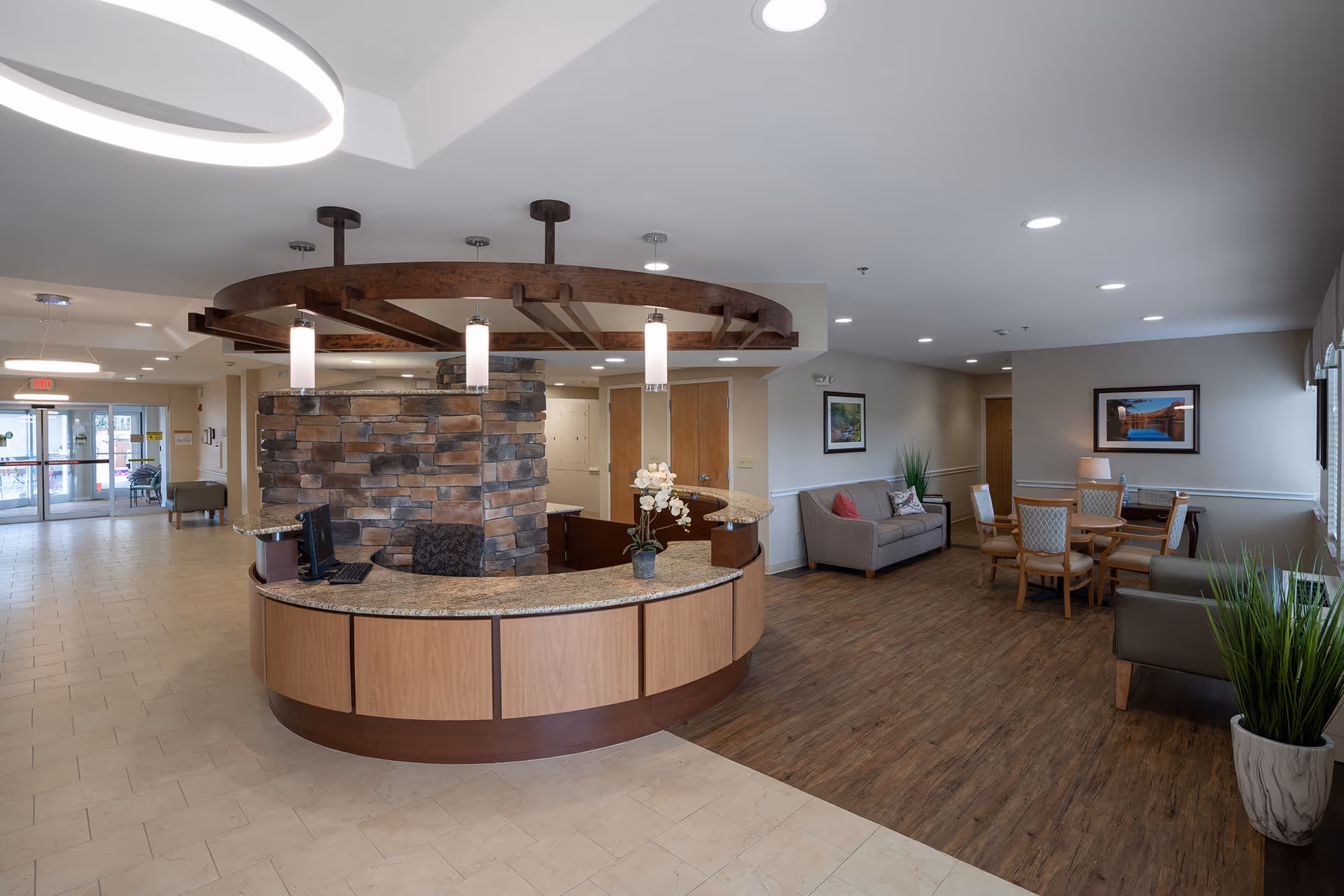 Curved reception desk with stone accent wall in a spacious assisted living lobby featuring seating and a dining area.