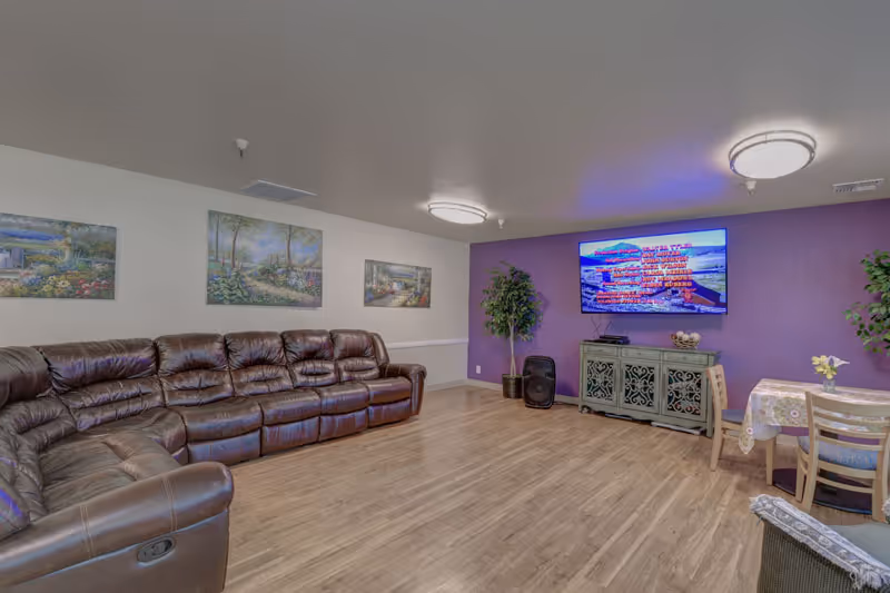 Spacious communal lounge with a large brown leather sectional, wall-mounted TV on a purple accent wall, a small dining table, and decorative plants and paintings.