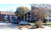 Brick two-story building with parked cars, small trees, and landscaping in a paved parking lot.