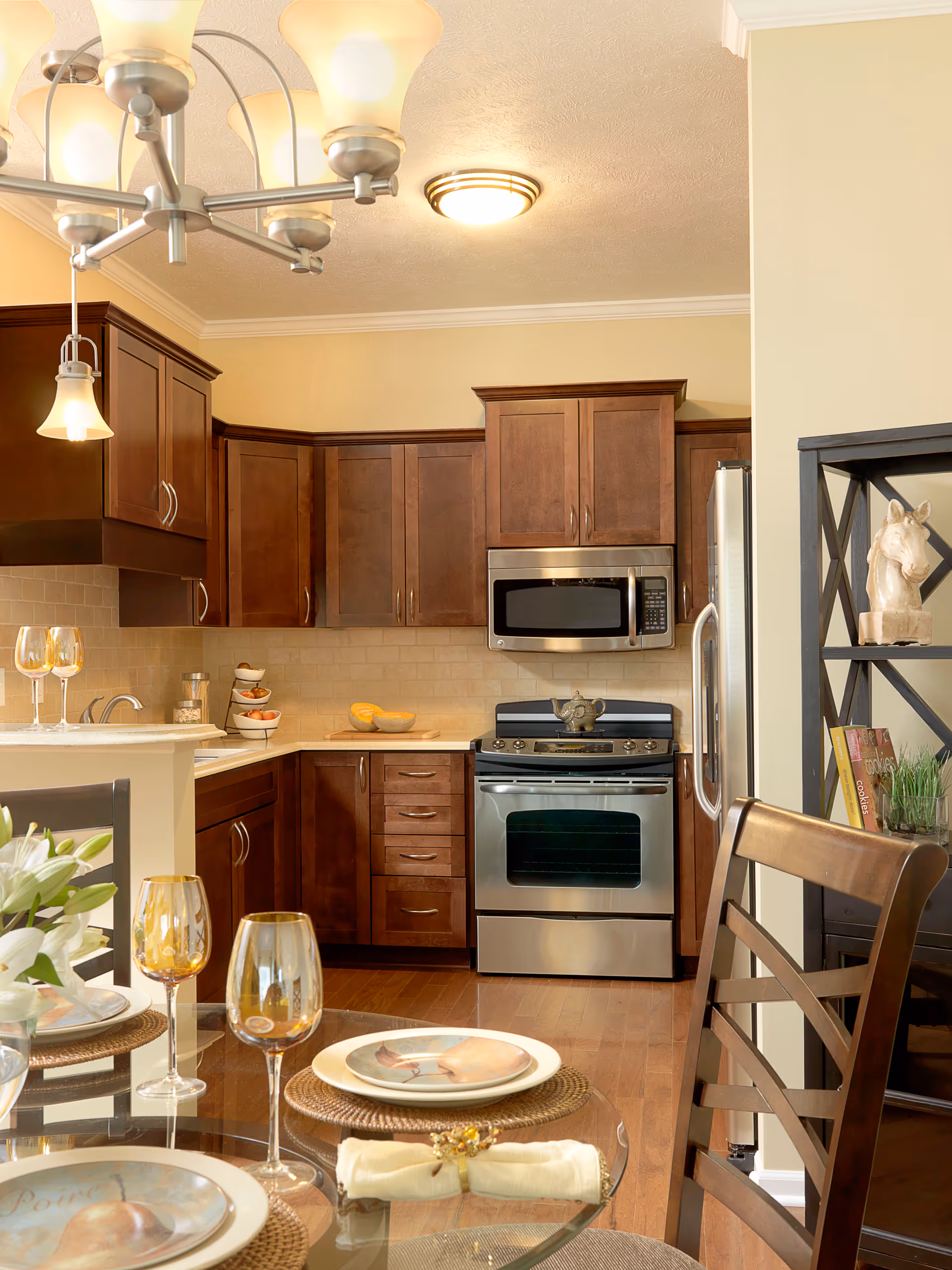 A modern kitchen with dark wooden cabinets, stainless steel appliances including a microwave, stove, and refrigerator. In the foreground, there is a glass dining table set with plates, napkins, and amber-colored wine glasses. A decorative shelving unit with books and a horse head sculpture is visible on the right side.