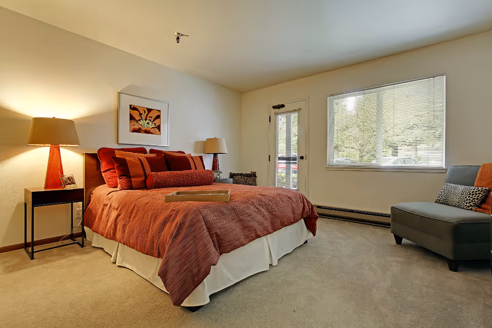 A cozy bedroom with a large bed covered in rust-colored bedding and multiple pillows. There are two bedside tables with lamps on either side of the bed. A framed floral artwork hangs above the bed. A gray armchair with a leopard print pillow and an orange pillow is positioned near a window with blinds. A door with a window leads outside, and the room is carpeted with beige flooring.