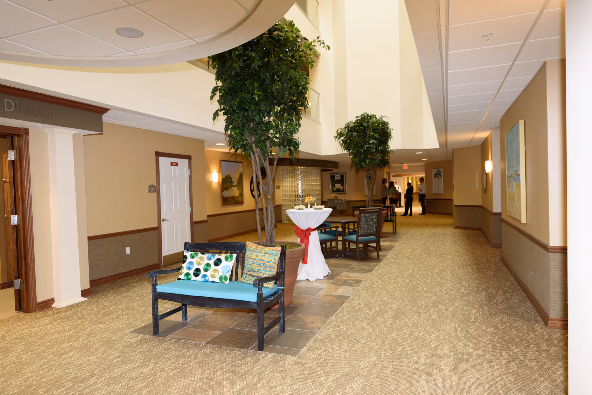 A spacious hallway in a senior living facility with beige walls and carpeted floors. The hallway features potted indoor trees, a small bench with colorful cushions, a tall round table draped with a white cloth and red ribbon, and several chairs and tables further down the corridor. There are paintings on the walls and a few people standing and conversing in the distance.