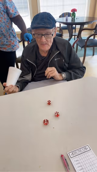 An elderly man wearing a blue cap and glasses is sitting at a table with three red dice in front of him. He is holding a white cup in one hand and appears to be playing a game called Bunco, as indicated by a score sheet and pen on the table. A person wearing a colorful patterned shirt and blue gloves is standing behind him. In the background, there are round tables and chairs near windows with blinds.