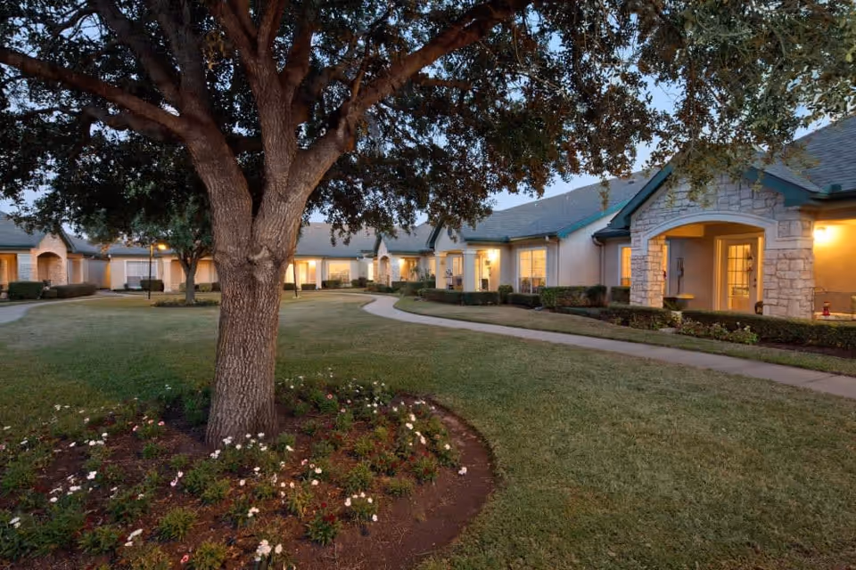 A landscaped outdoor courtyard area at dusk with a large tree surrounded by flowers in the foreground, a curved concrete pathway, and single-story buildings with lit windows and stone accents in the background.