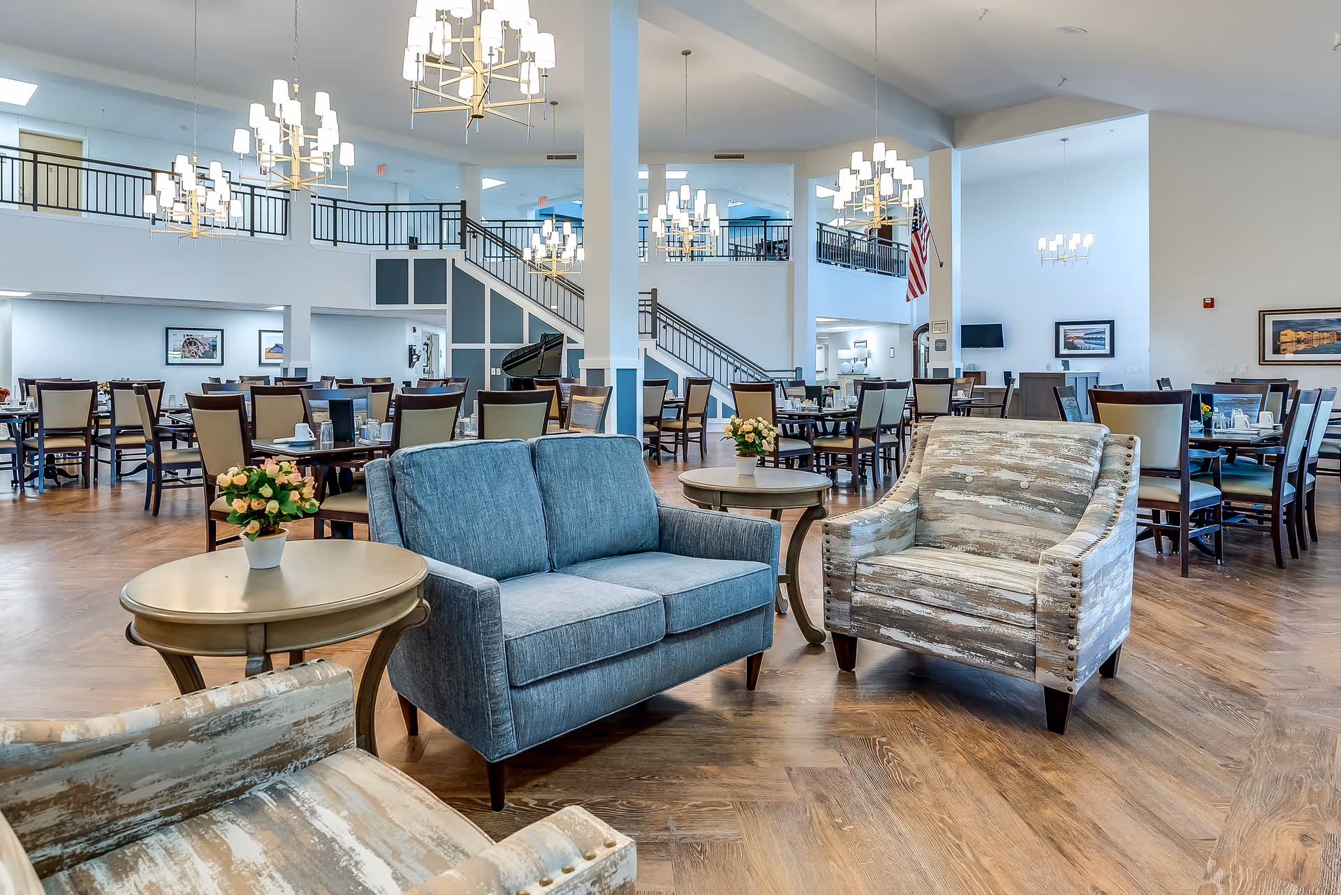 Spacious open senior living dining and lounge area with sofas, armchairs, multiple dining tables, chandeliers, and a central staircase.