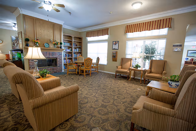 A cozy living room area in an assisted living facility with striped upholstered armchairs arranged around a carpeted floor. There is a brick fireplace with a wooden mantle, built-in bookshelves, a small table with chairs near the windows, and lamps providing warm lighting. The windows have striped valances and there are plants and framed pictures on the walls.