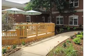 Curved concrete pathway leads past landscaped beds to a wooden deck with umbrellas in a courtyard beside a brick building.