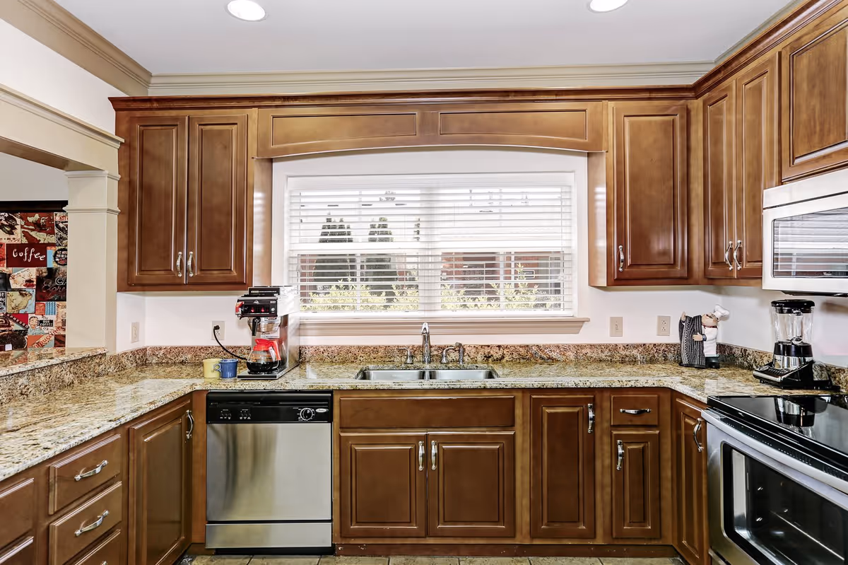 A kitchen with wooden cabinets, granite countertops, and stainless steel appliances including a dishwasher and oven. There is a double sink under a window with white blinds. On the countertop, there is a coffee maker, a blender, and a small chef figurine. The kitchen has recessed lighting in the ceiling.