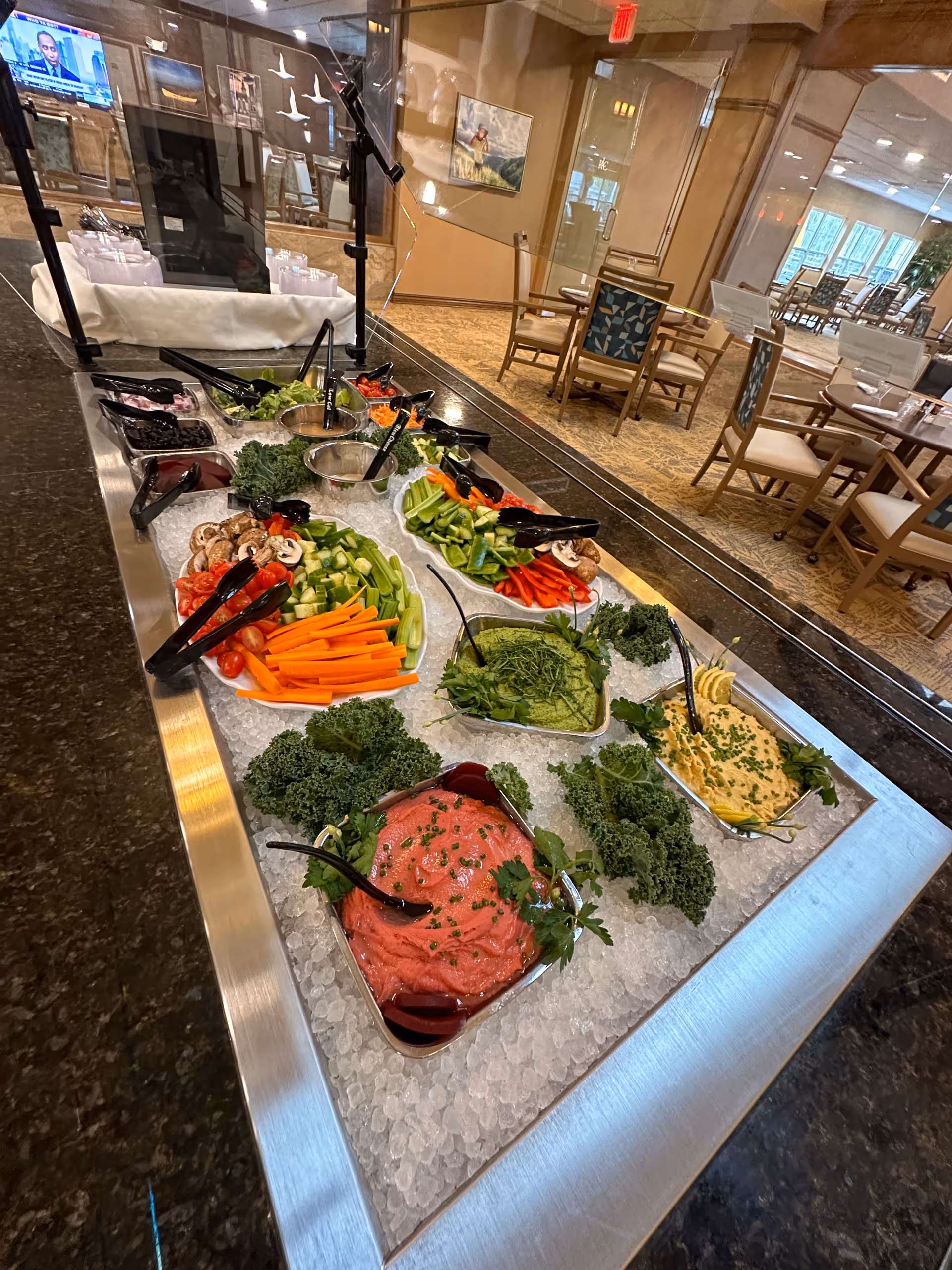 A salad bar with various fresh vegetables and dips displayed on a bed of ice, including carrots, celery, bell peppers, mushrooms, and leafy greens. The salad bar is situated in a dining area with tables and chairs visible in the background, separated by a glass partition.
