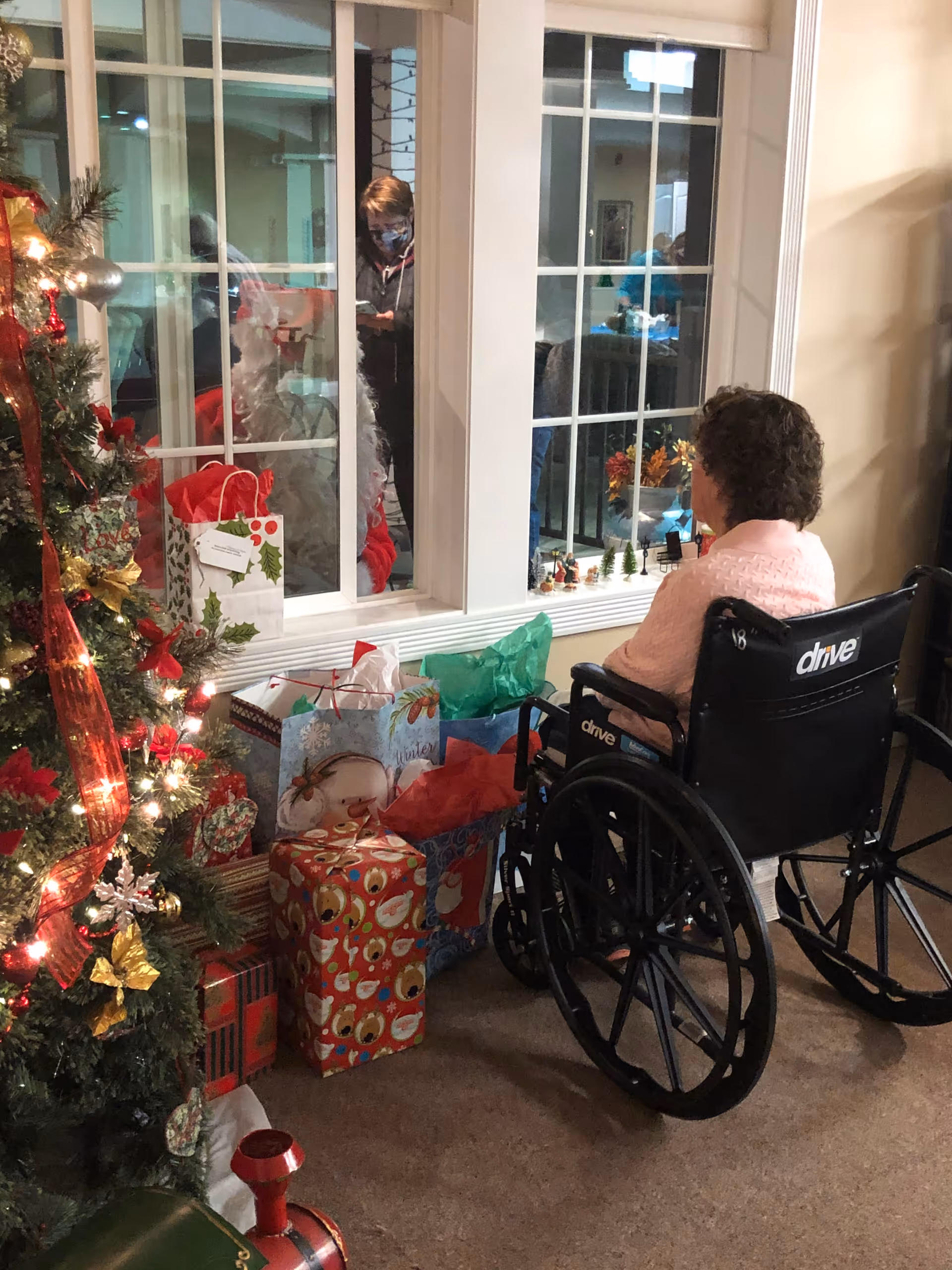 An elderly woman in a wheelchair wearing a pink sweater sits indoors near a decorated Christmas tree with wrapped presents underneath. She is looking out a large window where a person wearing a mask and dark clothing is visible on the other side. The room is warmly lit and decorated for the holiday season.