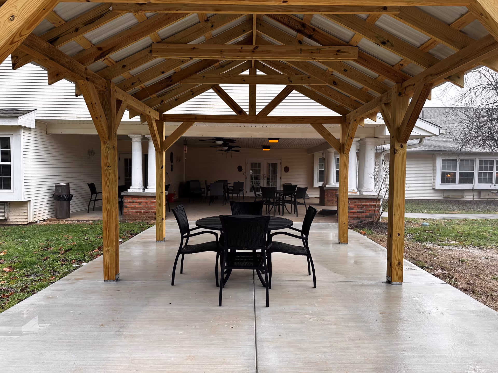 Outdoor covered patio area with a wooden roof structure and several black chairs and tables on a concrete floor. The patio is adjacent to a white building with columns and windows, and there is grass and some bare soil around the patio.