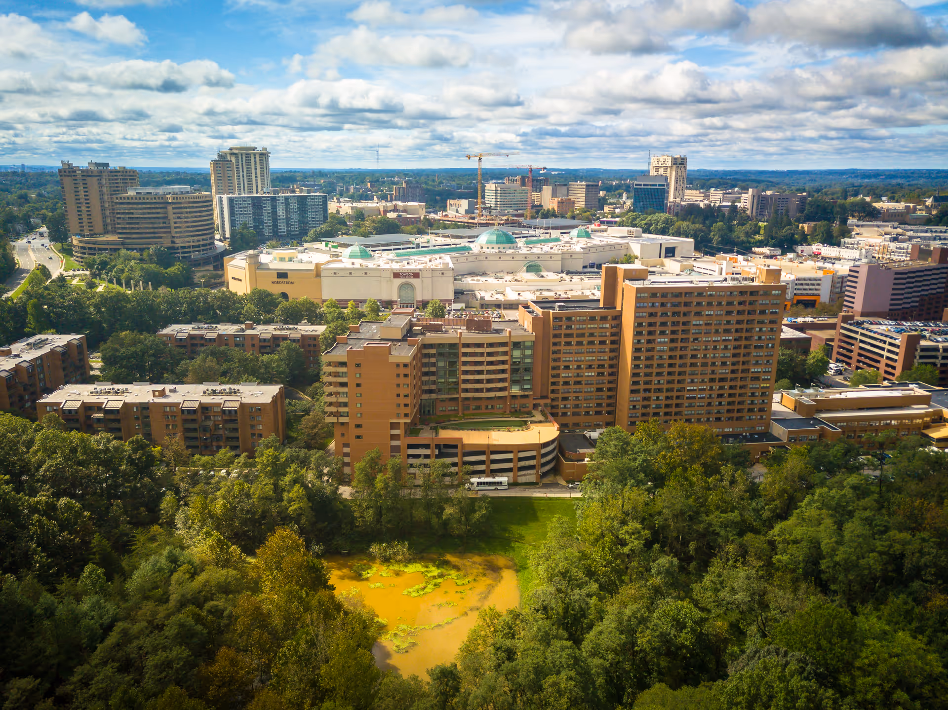 Aerial view of a cityscape featuring multiple tall buildings, including residential and commercial structures, surrounded by dense green trees and a small pond in the foreground under a partly cloudy sky.