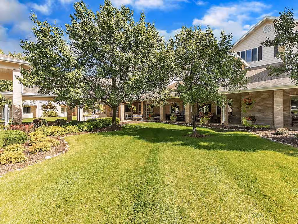 A well-maintained outdoor garden area at Holiday Genesee Gardens featuring green grass, two leafy trees, and a building with a covered porch and hanging flower baskets under a partly cloudy blue sky.