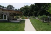 Concrete walkway through a lawn leading to a wooden porch/pavilion and a fenced garden with trees in the background.