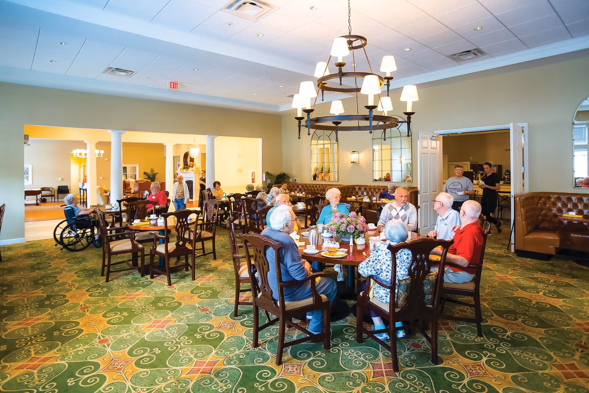 A spacious dining room in a senior living facility with elderly residents sitting around tables, eating and chatting. The room features a patterned green carpet, wooden chairs, a large chandelier with multiple lampshades, and mirrors on the wall. There are staff members in the background attending to the residents.
