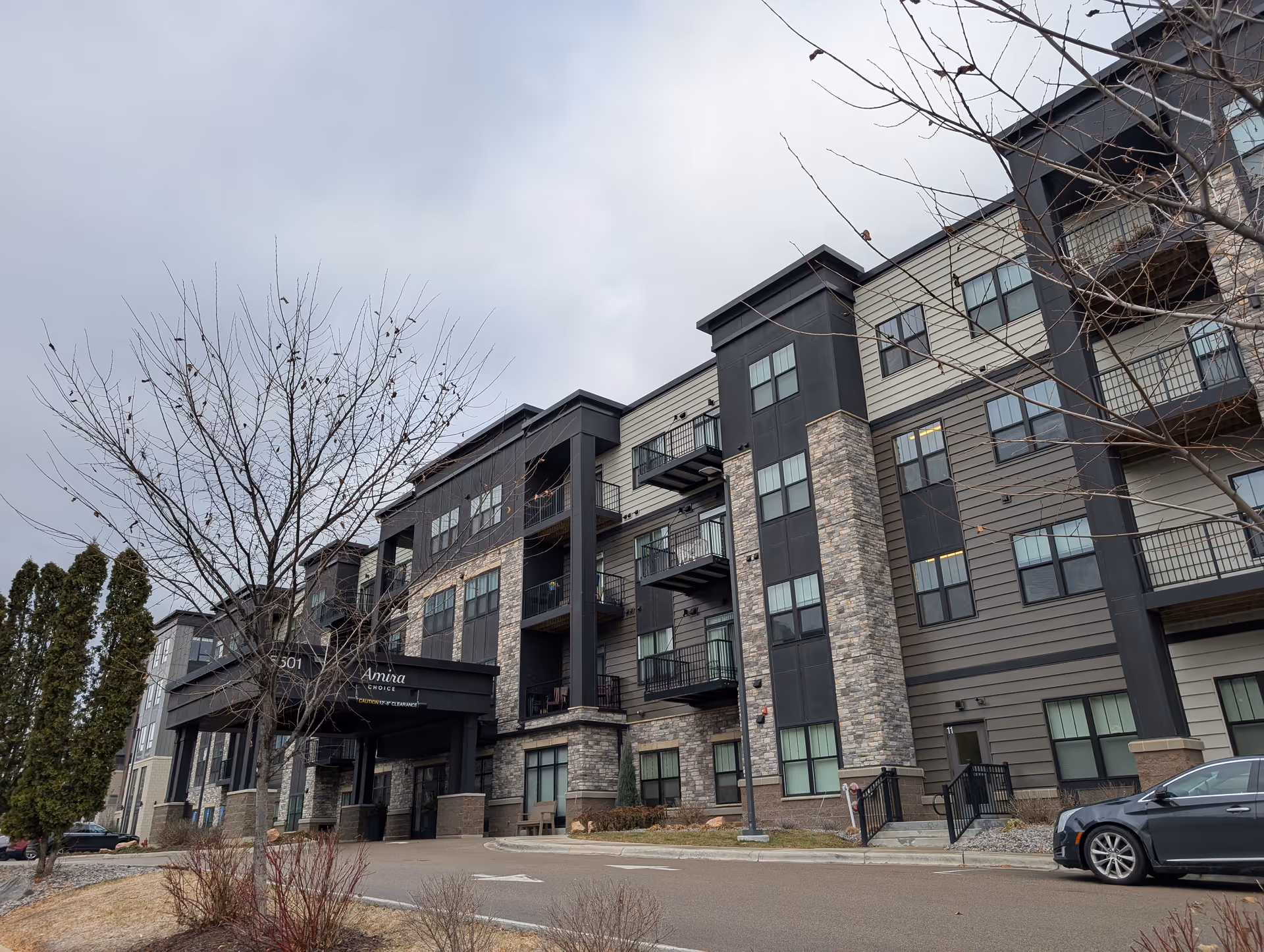 Exterior view of a multi-story senior living facility named Amira Choice Bloomington, featuring a modern design with stone and siding facade, balconies, and a covered entrance. The sky is overcast and there are leafless trees and some shrubs in the foreground.