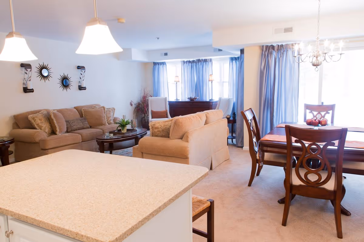 A bright and spacious living and dining area with beige sofas, a wooden coffee table, and a dining table with four chairs. The room has large windows with blue curtains allowing natural light to fill the space. Decorative wall mirrors and lamps add to the cozy ambiance.