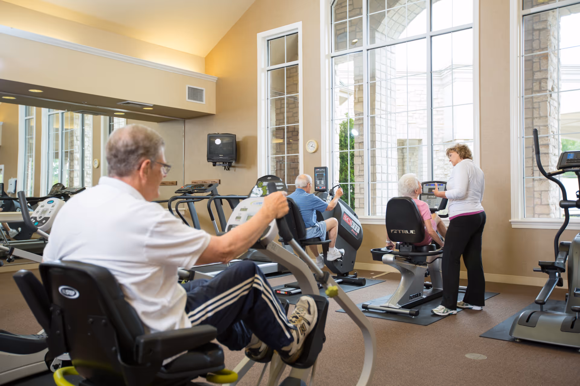 An indoor fitness room with large windows letting in natural light. Several senior adults are using exercise equipment, including stationary bikes and elliptical machines. A fitness instructor is assisting one of the seniors. The room has beige walls, a large mirror, and exercise machines arranged neatly.