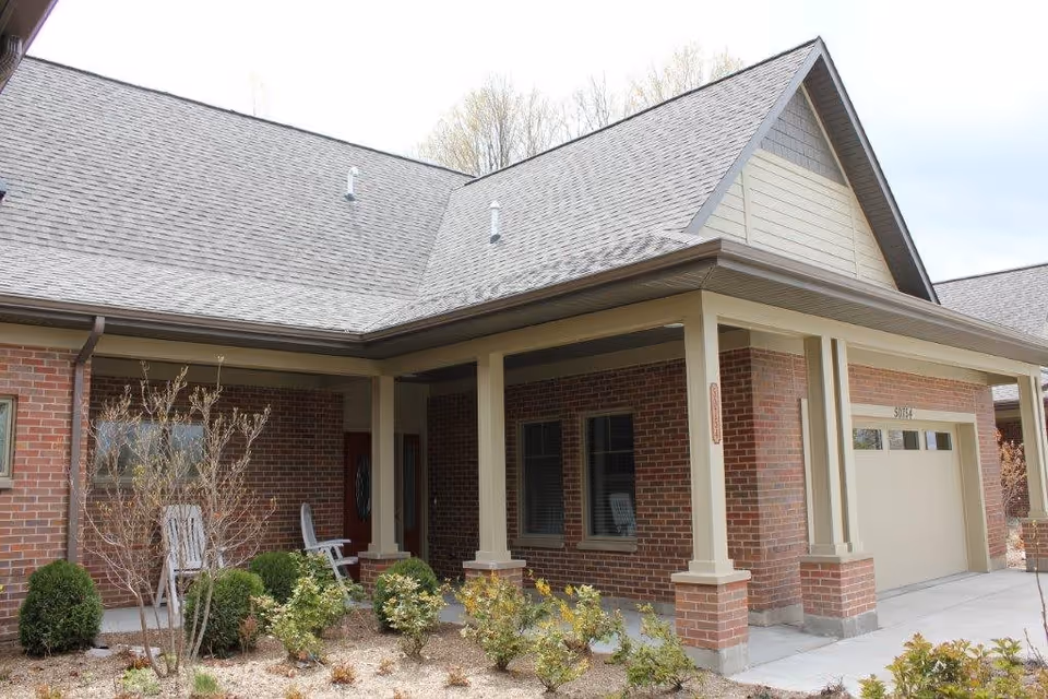Exterior view of a brick building with a covered porch supported by columns, a garage door, and some landscaping with bushes and small trees in front.