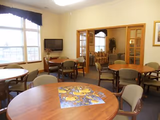 A common area with multiple round wooden tables and chairs arranged around them. A large window with dark valance allows natural light into the room. A flat-screen TV is mounted on the wall near a small table with a decorative plant. In the background, a seating area with sofas is visible through glass-paned double doors.
