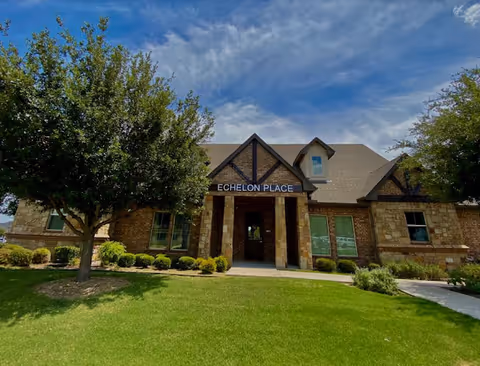 Front view of a stone and brick assisted living building with a covered entrance labeled 'Echelon Place', surrounded by a lawn and trees.