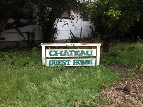 Overgrown front yard with weathered Chateau Guest Home sign in front of a house.