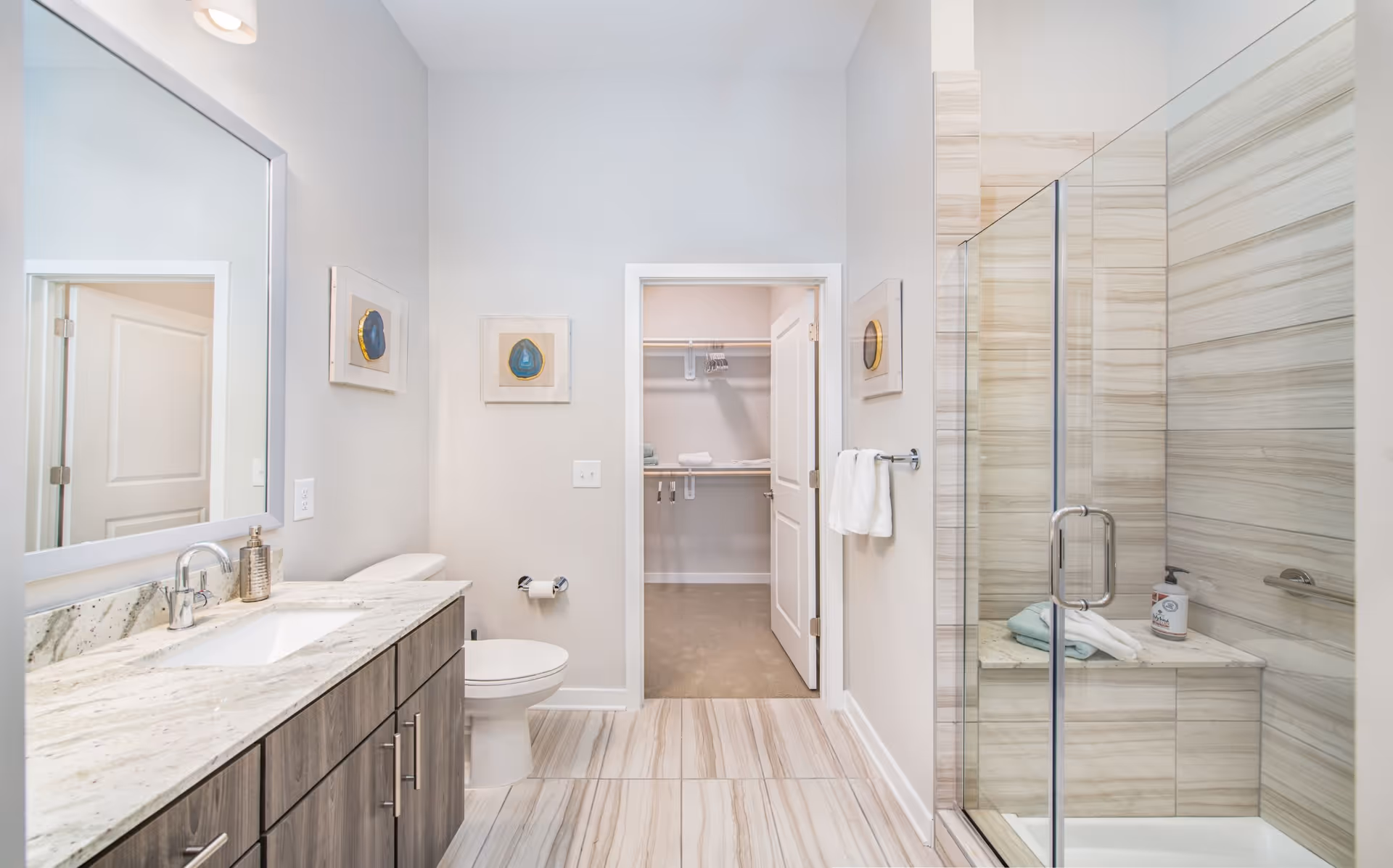 Bright and modern bathroom with a large mirror above a granite countertop vanity, a toilet, and a glass-enclosed shower with beige tile walls. The bathroom has light-colored walls and floor tiles, two framed artworks on the walls, and an open door leading to a walk-in closet with shelves and hangers.
