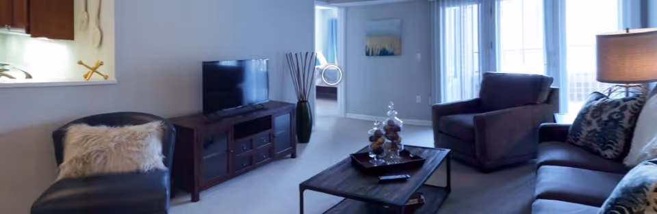 A cozy living room with a dark leather chair with a fluffy pillow, a wooden TV stand with a flat-screen TV, a coffee table with decorative jars, a dark armchair, and a sofa with patterned cushions. The room has light-colored walls and carpet, with a lamp on a side table and a window with vertical blinds letting in natural light.