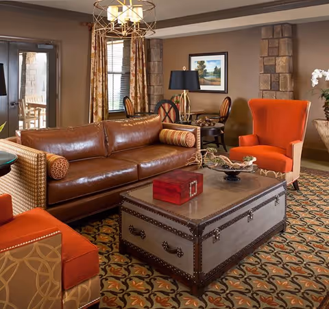 A cozy living room area with a brown leather sofa, two orange armchairs, a large decorative trunk used as a coffee table, and a patterned carpet. The room features stone pillars, a framed landscape painting on the wall, a table with chairs in the background, and a door leading outside.