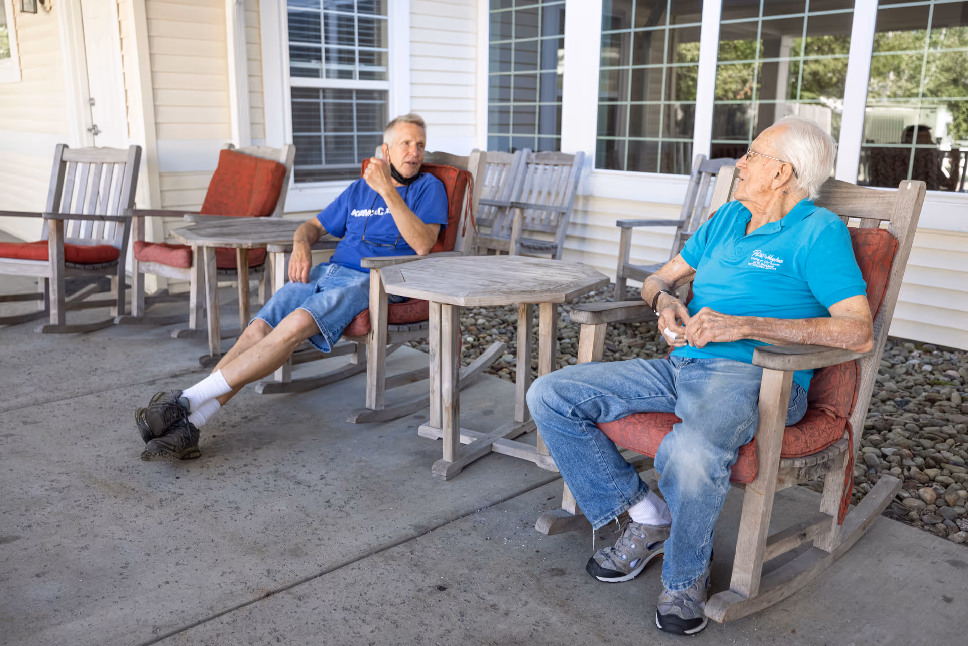 Two elderly men sitting and conversing on wooden rocking chairs with red cushions on a covered patio outside a building with large windows.