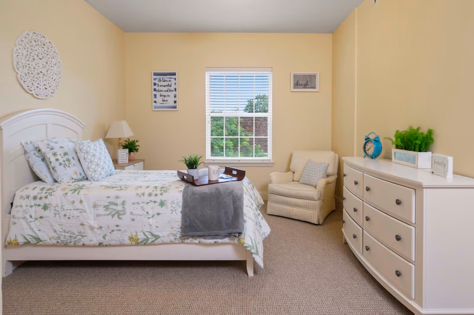 A cozy bedroom with a white bed frame and floral bedding. There is a wooden tray with a mug and a small plant on the bed. A beige armchair with a pillow is placed near a window with white blinds. A white dresser with drawers holds a blue alarm clock, a rectangular planter with greenery, and a small sign that says 'family'. The walls are painted light yellow and decorated with framed quotes and a white decorative wall piece.